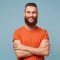 Studio portrait of emotional happy funny smiling boyfriend man with a heavy beard stands with arms crossed dressed in red t-shirt isolated over blue background Studio portrait of emotional happy funny smiling boyfriend man with a heavy beard stands with arms crossed dressed in red t-shirt isolated over blue background